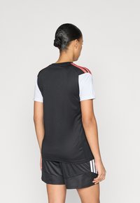 Μαύρη αθλητική φανέλα με λευκά κοντά μανίκια, τρεις κόκκινες ρίγες στους ώμους και αντιστοιχισμένο μαύρο σορτς με λευκές ρίγες στα πλάγια.