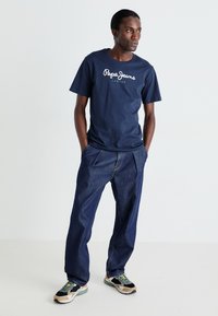 Jūrinės medvilnės marškinėliai su balta „Pepe Jeans London“ logotipu, derinami su tamsiai mėlynomis džinsinėmis kelnėmis su klostėmis priekyje ir kasdieniais sportbačiais.