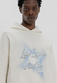 Kremase hoodie med frontlomme, med metallisk tekst "Low Lights Studios" og blå airbrush-akenter. Myk stofftekstur.