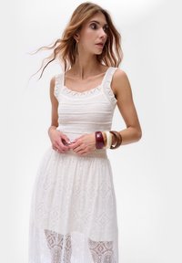 Débardeur en crochet blanc avec un décolleté carré, associé à une jupe en dentelle blanche transparente présentant un design à motifs et une taille ajustée.