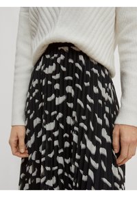 Suéter blanco acanalado metido en una falda negra con pliegues y patrones abstractos de pinceladas blancas y grises, con las manos descansando a los lados.