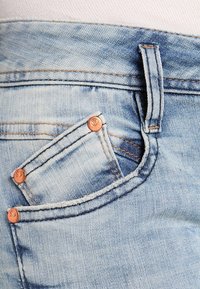 Světle modré denimové džíny s viditelným šitím, malou přední kapsou a měděnými nýty, které vyzdvihují design.