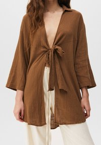 Femme portant une chemise marron à texture lâche nouée à l'avant avec de larges manches et un pantalon couleur crème.