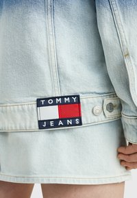 Hele sinise denimburger, millel on Tommy Jeansi tempel, hõbedased metalist nupud ja toores serv. Kangas omab pleekinud ja tekstuurset välimust.