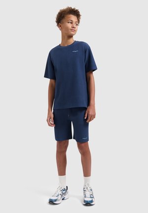 Tienerjongen staat, draagt een bijpassend navy blauw geribd T-shirt en korte broek met witte sokken en blauw-witte sneakers op een effen achtergrond.