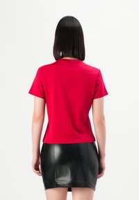 Donna con capelli neri lisci, che indossa una maglietta rossa a maniche corte e una minigonna di pelle nera, voltata di spalle contro uno sfondo bianco uniforme.
