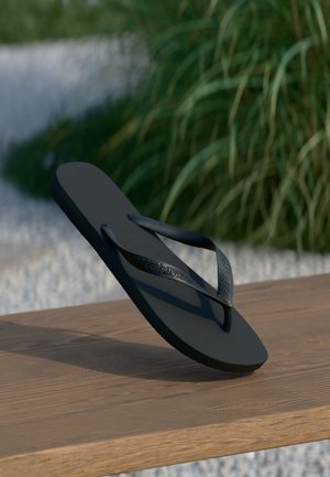 Black rubber flip-flop with textured straps on wooden plank, green leaves and white gravel blurred in background.