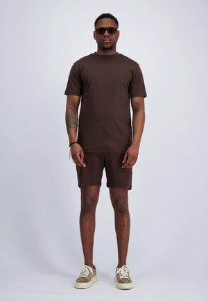 Mann trägt dunkelbraunes T-Shirt und passende Shorts, beige Sneakers, Sonnenbrille und ein Tattoo auf dem linken Unterarm, steht vor einfarbigem grauen Hintergrund.