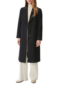Manteau noir sur mesure à coupe droite, doté d'un col cranté et de trois boutons. Porté sur un haut beige clair et un pantalon ample crème.