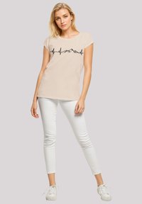 Beige cap-sleeve top with a heartbeat line graphic; paired with white skinny jeans and white sneakers. Smooth fabric, casual design.