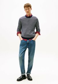Giovane uomo in piedi con le mani in tasca, indossa un maglione grigio a fantasia sopra una camicia a righe rossa, jeans blu e scarpe nere.