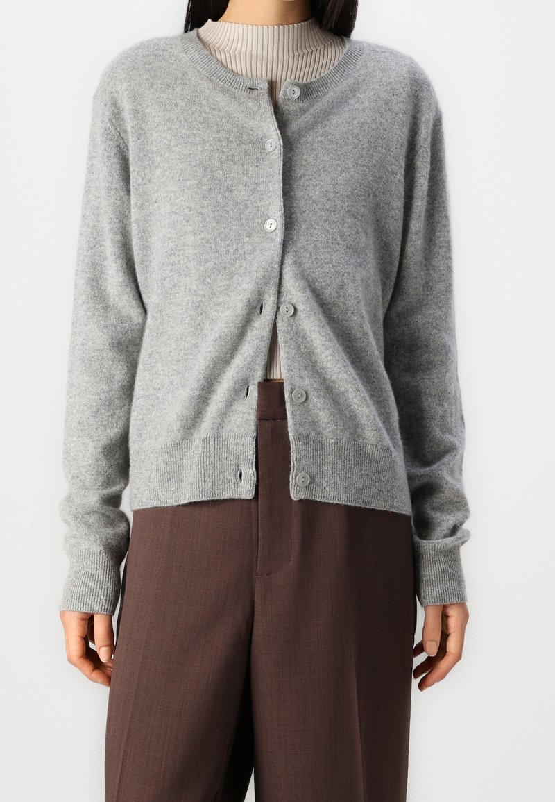 Femme portant un cardigan gris clair boutonné sur un top côtelé beige associé à un pantalon marron à taille haute, sur un fond uni.