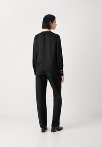 Blouse noire à manches longues avec un motif texturé, des manches froncées et des accents de boutons. Associée à un pantalon noir et des chaussures à talons.