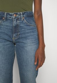 Jeans blu a vita alta con lavaggio sbiadito, chiusura con bottone metallico, tasche anteriori e dettagli di cucitura. Design slim fit ben visibile.