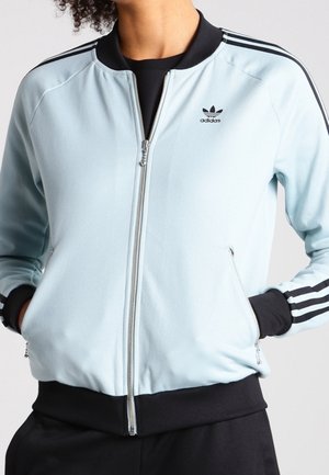 Personne portant une veste zippée Adidas bleu clair avec col, poignets et ceinture noirs, ainsi que trois bandes noires sur les manches.