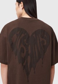 Brun bomulds-t-shirt med stort sort dryppende hjertegrafik på ryggen. Oversized pasform med korte ærmer og rund halsudskæring.