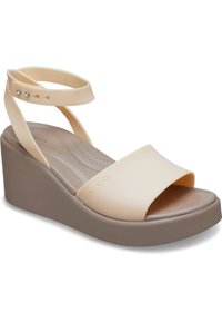 Sandales à talon compensé avec une tige beige clair et une semelle en caoutchouc. Possède une large sangle et des lanières de cheville, texture lisse, avec un détail du logo "crocs".