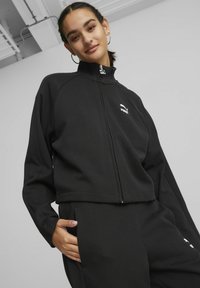 Schwarze Zip-Jacke mit hohem Kragen und langen Ärmeln. Mit einem weißen Puma-Logo und einer strukturierten Oberfläche. Kombiniert mit passenden Hosen.