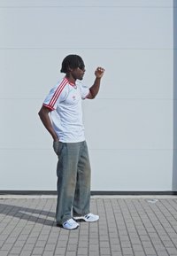 Vit sporttröja med röda ränder, lösa grå jeans och vita sneakers med svarta detaljer. Stående pose mot en ljus bakgrund.