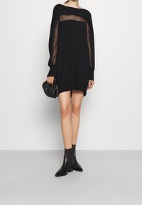 Robe noire ample avec des panneaux en dentelle sur les manches, associée à un petit sac à main noir et des bottines noires. Texture lisse, coupe décontractée.