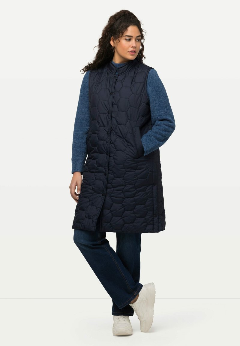 Lång marinblå vadderad väst med hexagonmönster, utrustad med ståkrage, framknappar och sidofickor, tillsammans med en blå tröja och jeans.