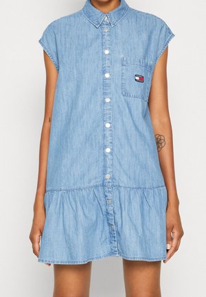 Ærmeløs lyseblå denimskjortekjole med knapper foran, brystlomme med logo og volanthæm, båret af person med arm tatoveringer.