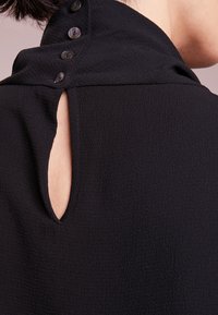 Närbild av en svart strukturerad blus baksidan med en liten nyckelhålsöppning och fyra svarta knappar längs kragen.