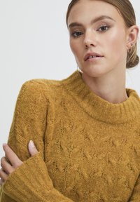 Senfgelbes Strickpullover mit hohem, geripptem Kragen, strukturiertem Diamantmuster und breiten Bündchen. Weiche, kuschelige Textur ist sichtbar.