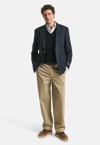 Marine gestreepte blazer over een marineblauwe trui, wit geruit shirt, beige broek en bruine schoenen. Kenmerkt knopen en een zak op de blazer.
