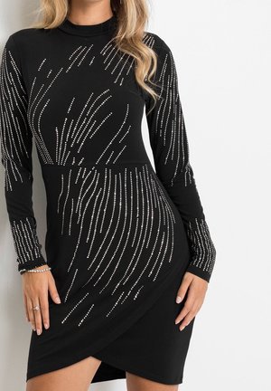 Femme portant une robe noire ajustée à manches longues avec des motifs en strass argentés en forme de rayures et un ourlet de jupe superposé.