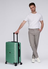 Hombre con camiseta blanca y pantalones grises de pie, sosteniendo el asa de una gran maleta verde con ruedas sobre un fondo blanco.
