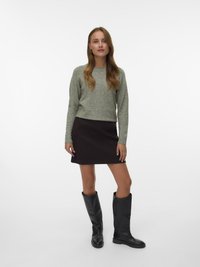Una donna in piedi indossa un maglione grigio a maglia, una mini gonna marrone scuro e stivali neri fino al ginocchio, di fronte a uno sfondo bianco semplice.
