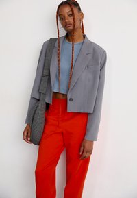 Jonge vrouw met lange rode vlechten, draagt oorringen, een grijze cropped blazer over een blauwe crop top, rode broeken en een geruite tas.