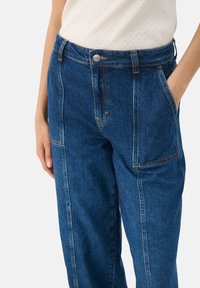 Jeans i mörkblå denim med kontrasterande orange sömmar, hög midja och sidofickor. Har en metalldetalj med knäppning.