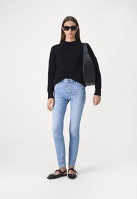 Suéter negro de punto, jeans de cintura alta azul claro con detalles de botones, zapatos planos negros y gafas de sol negras de gran tamaño. Diseño simple y ajustado.
