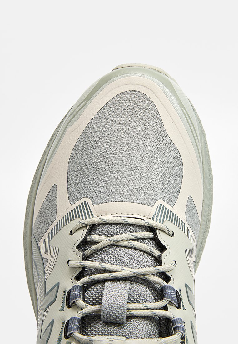 Gros plan sur une chaussure de sport grise et beige avec une tige en mesh, une languette rembourrée et des lacets entrecroisés passés à travers des œillets en plastique.