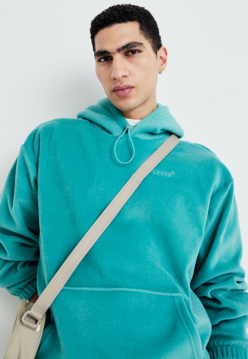 Türkiser Fleece-Hoodie mit Kordelzug, Fronttasche und gesticktem Logo. Getragen mit einer beigen Umhängetasche. Weiche, plüschige Textur.