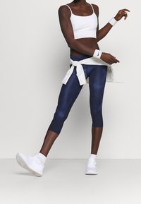 Marina sportleggings med en slät textur, vit kortärmad linne, och vita handledsstöd. Skorna är vita träningsskor i en avslappnad stil.