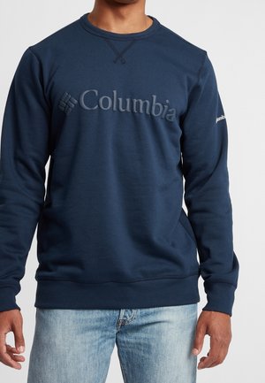 Mężczyzna w granatowej bluzie Columbia z logo na piersi i jasnoniebieskich jeansach na jednolitym tle.