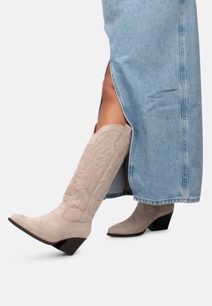 Nogi w jasnobeżowych, haftowanych kowbojskich kozakach do kolan oraz niebieskiej spódnicy dżinsowej z rozcięciem, jedna noga uniesiona, ukazując szczegóły buta.