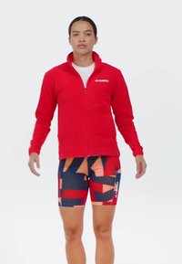 Rote, aufknöpfbare Sportjacke mit hohem Kragen, kombiniert mit gemusterten Shorts, die geometrische Formen in Marineblau, Orange und Rot zeigen.