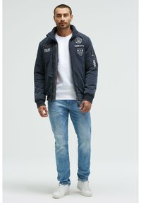 Marineblaues Bomberjackett mit Frontreißverschluss, gerippten Bündchen und mehreren Aufnähern. Getragen über einem weißen Shirt und kombiniert mit hellblauen Jeans und weißen Sneakers.