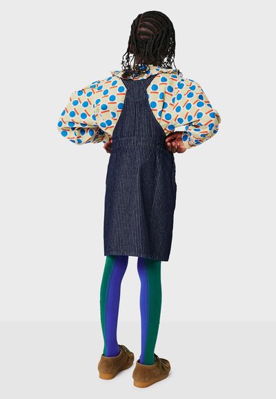 Salopette en denim à rayures verticales, superposée à un chemisier à motifs circulaires. Collants verts et bleus, chaussures marron à lacets.