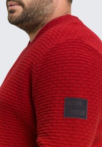Rode gebreide trui met een gestructureerd patroon, ronde halslijn en een zwart label op de linkermouw met merkinformatie.