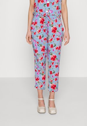 Femme portant un pantalon cropped bleu clair avec un imprimé floral rouge et rose, associé à des chaussures à plateforme Mary Jane blanches.