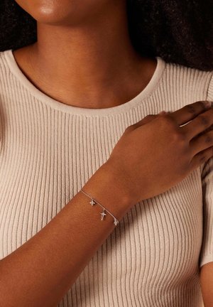 Bracelet en argent avec des breloques, y compris une étoile et des formes florales, porté sur un bras à peau foncée, devant un fond de haut beige à côtes.