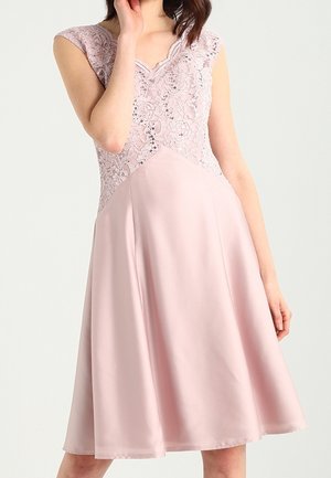 Vrouw draagt een mouwloze lichtroze jurk met kant- en pailletten details op het lijfje en een gladde, uitlopende rok.