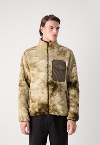 Faux fur jas in beige met een bruin spetterpatroon. Voorzien van een hoge kraag en een borstzak in olijfgroen met knoopsluiting. Voorkant met rits.