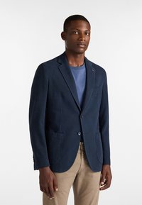 Mörkblå blazer i texturerad tyg, med enfaldig knäppning, notch-kragar och två framfickor, som bärs över en blå t-shirt.