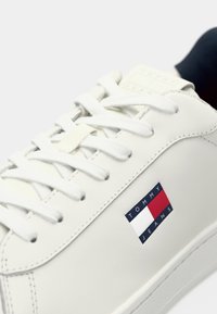 White sneaker with white laces featuring a red, white, and blue Tommy Jeans logo on the side and a black sock visible inside.